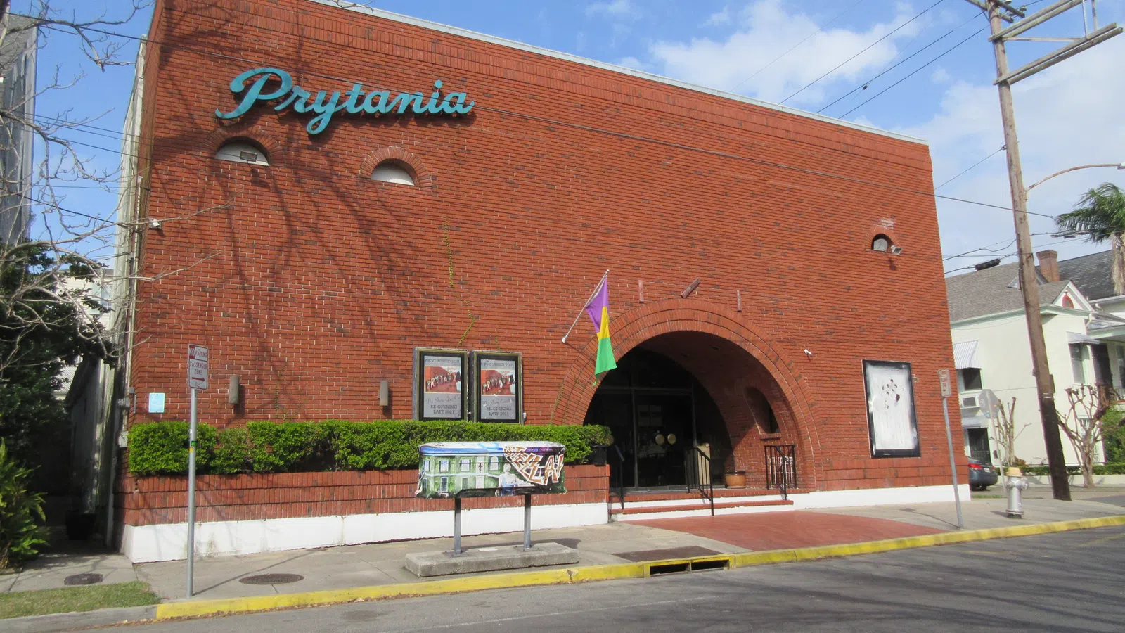Red brick building with arched entrance and Prytania sign under blue sky, surrounded by greenery and street elements.