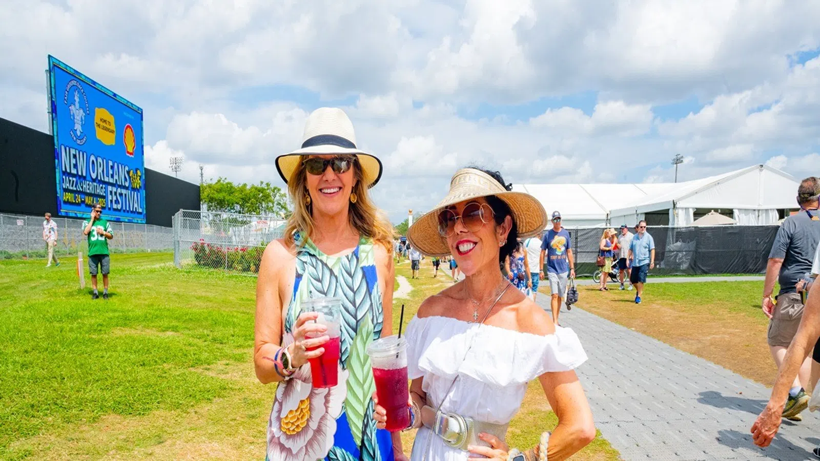 Two women in summer dresses and hats holding drinks at New Orleans Jazz Festival under bright sky.