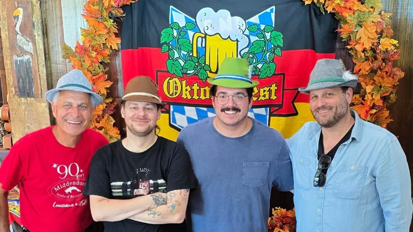 Four men wearing festive hats pose smiling in front of an Oktoberfest banner with autumn leaves decorations.
