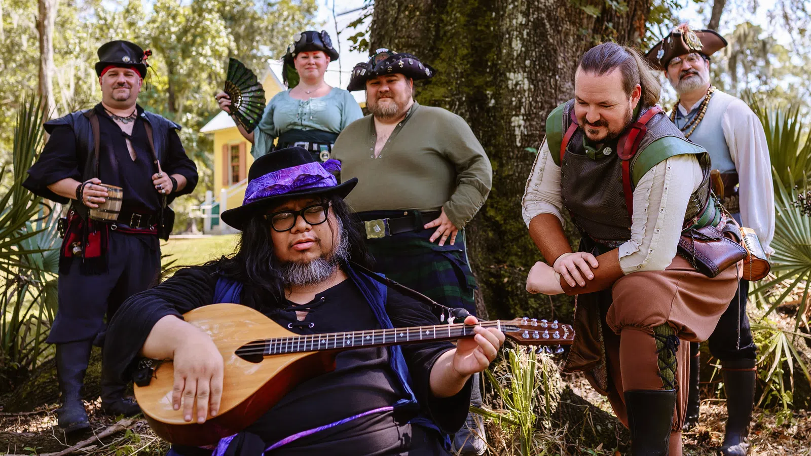 Group of six people in medieval and pirate costumes posing outdoors with a lute player in front under a large tree.