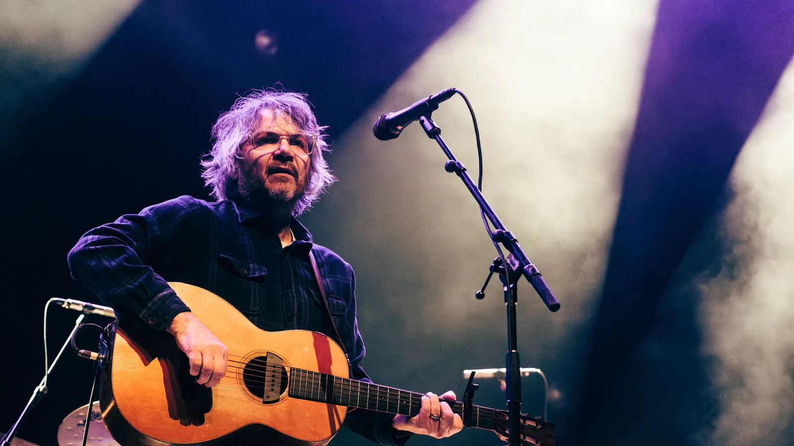 Musician playing acoustic guitar on stage with microphone and colorful stage lighting in smoky background.