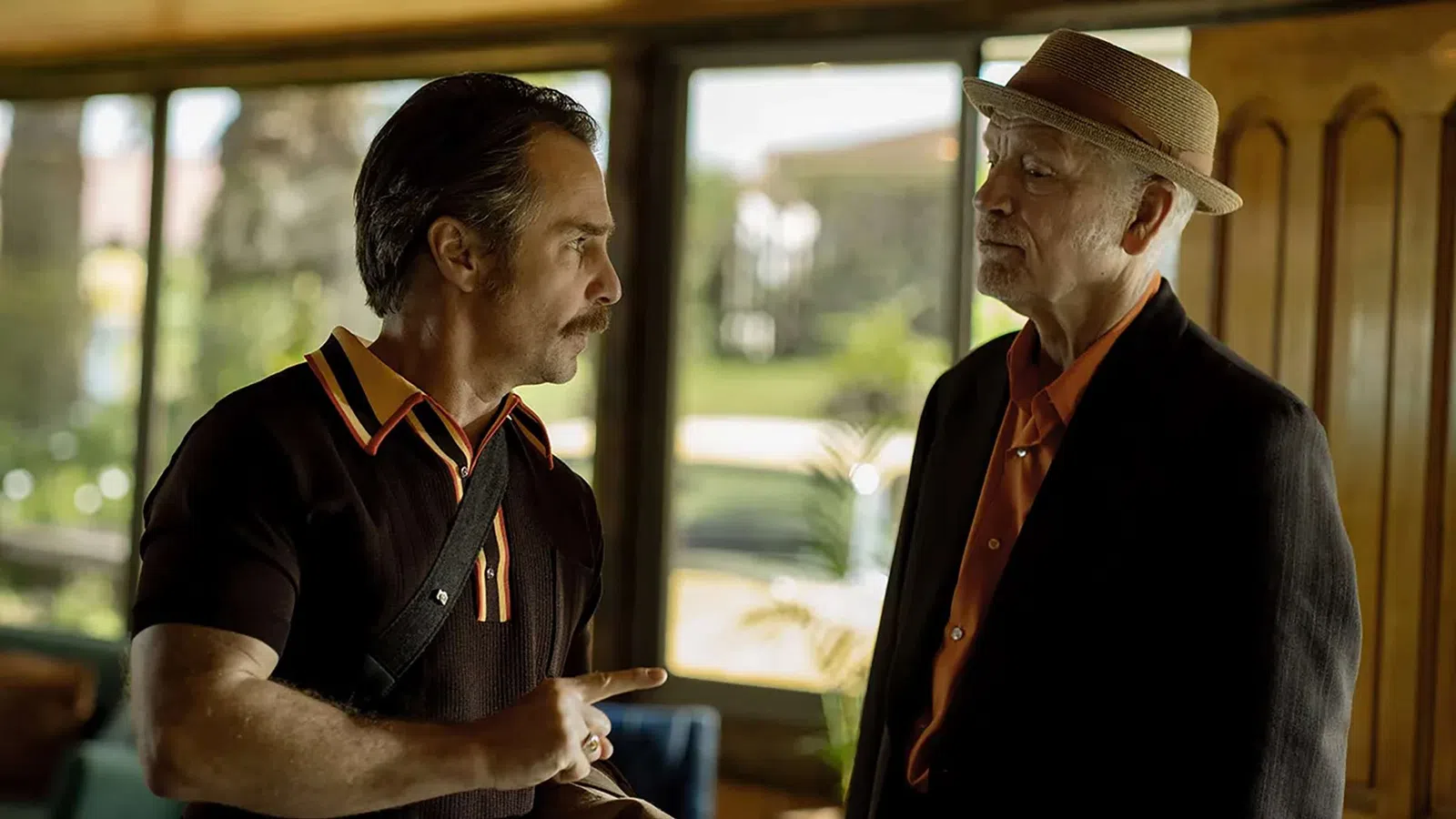 Two men in conversation, one wearing a hat and suit, the other in a black shirt with orange collar details indoors near windows.