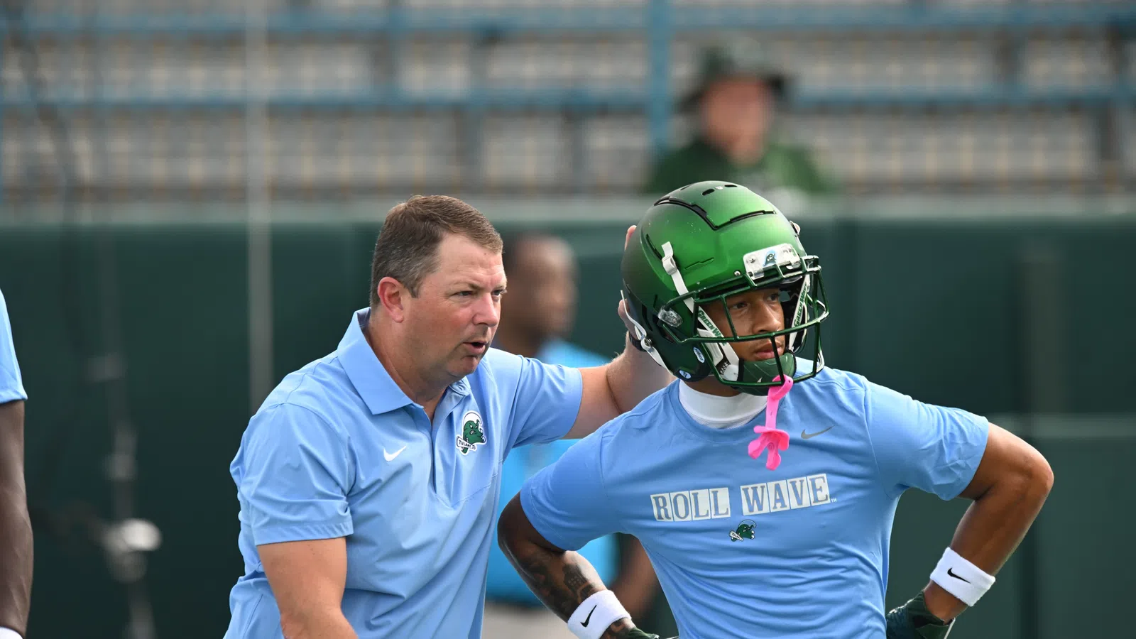 Football coach instructing player wearing green helmet and blue Roll Wave shirt during practice session