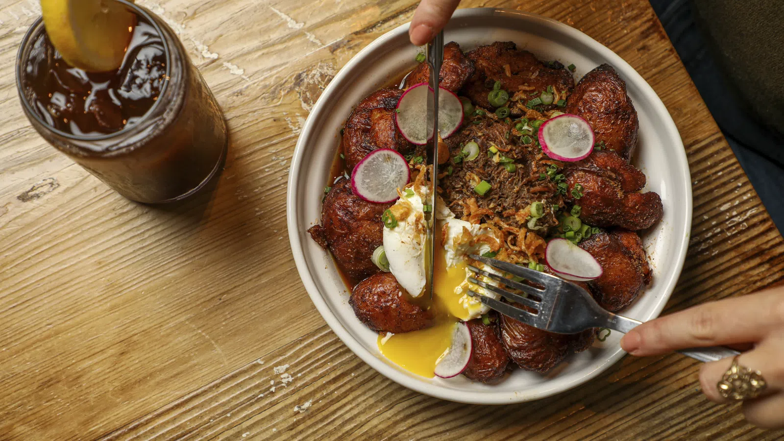 Bowl of fried plantains with poached egg, shredded meat, and radish slices on a wooden table with iced tea.