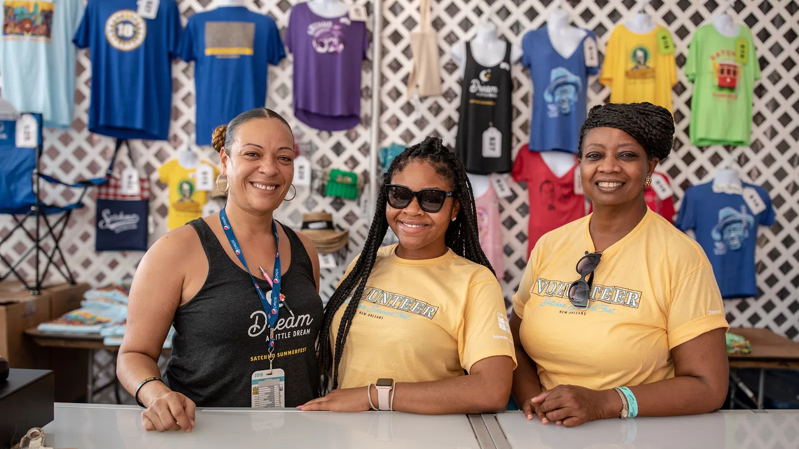 Three smiling women standing behind a counter with colorful t-shirts hanging on the wall behind them