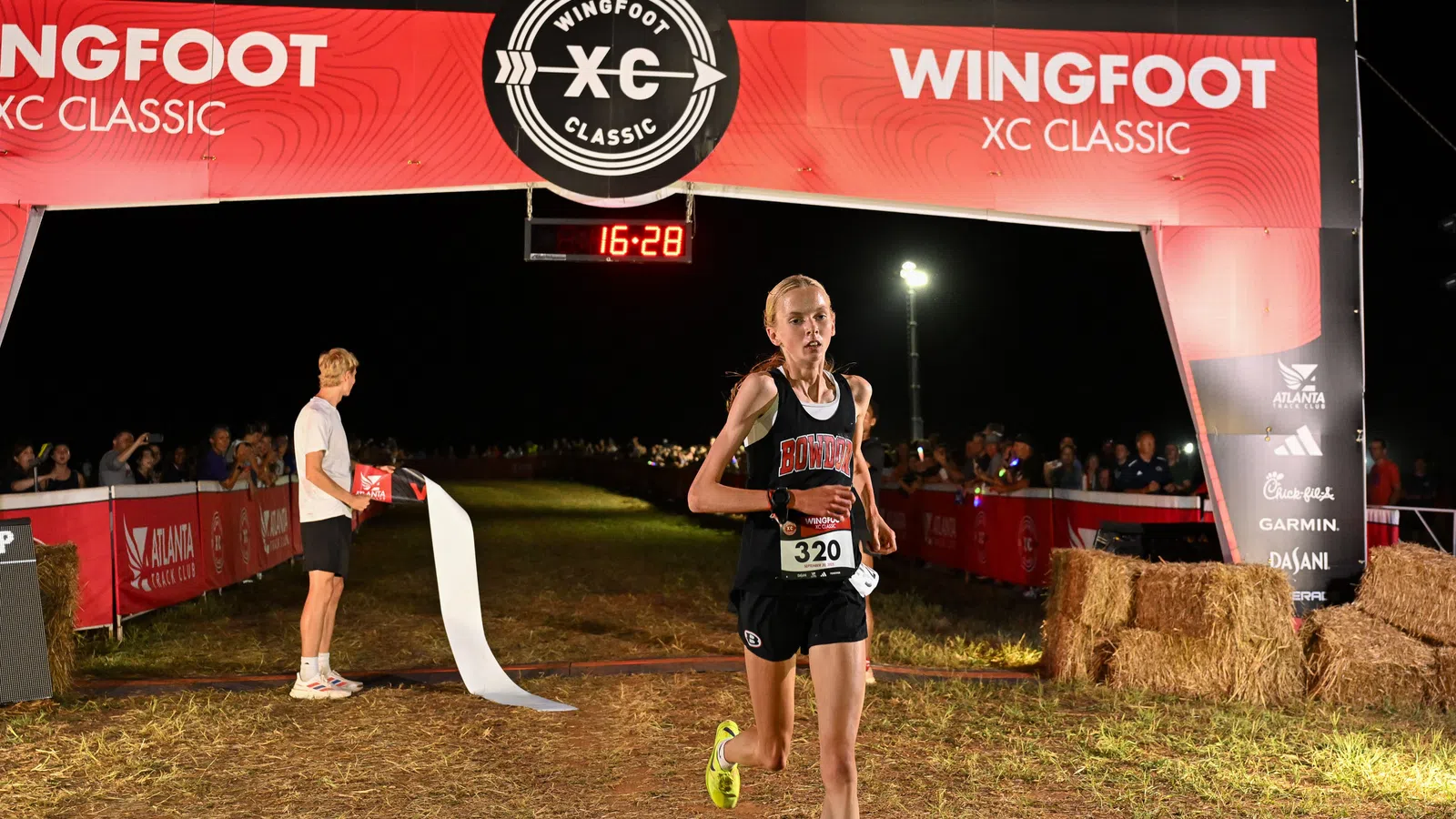 Female runner in black uniform crosses finish line at Wingfoot XC Classic nighttime race with crowd and timer visible.