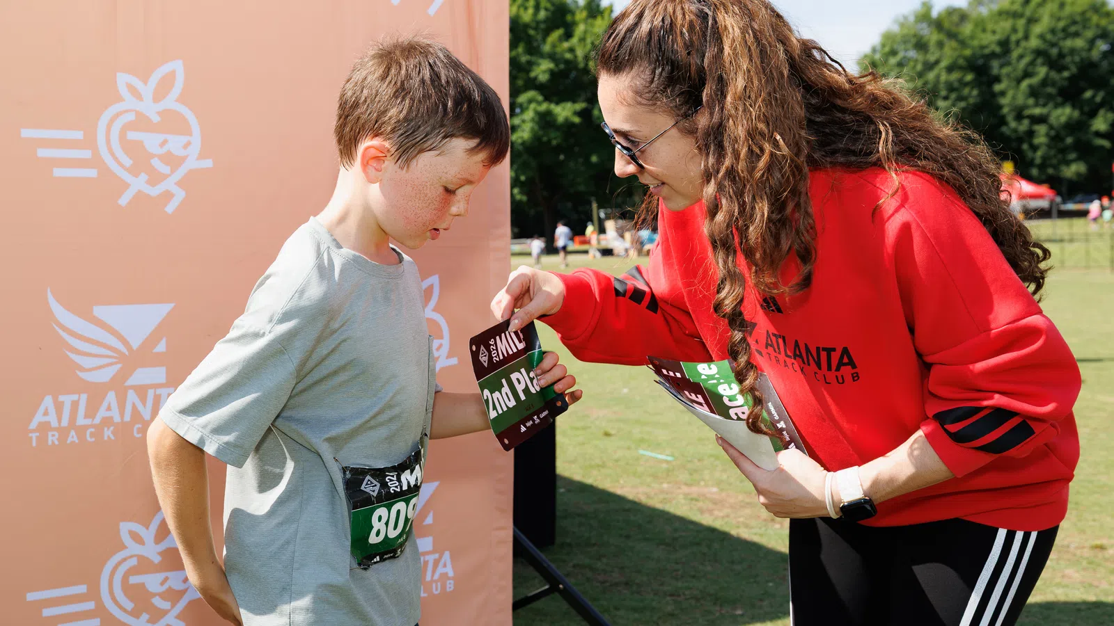 Young boy receiving 2nd place award from woman in red Atlanta Track Club sweatshirt at outdoor event.