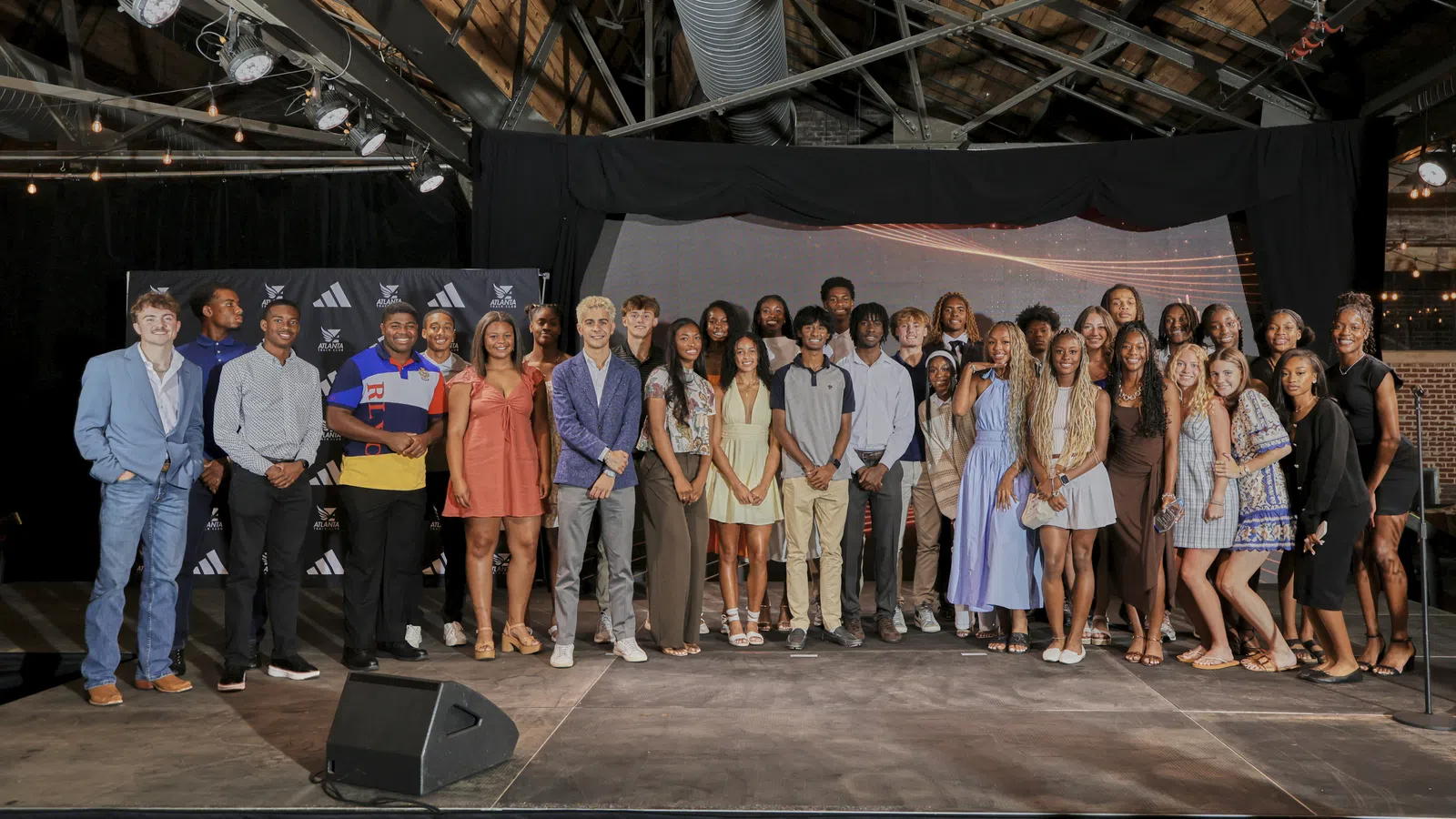 Large diverse group of young adults posing on a stage inside a rustic venue with wooden beams and lighting fixtures.