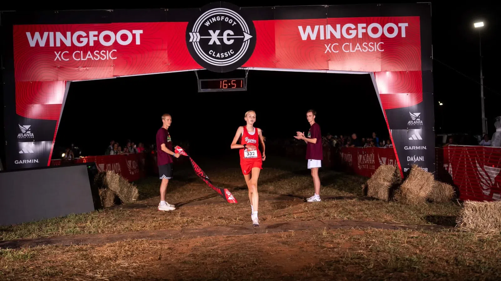 Female runner crosses finish line at Wingfoot XC Classic event at night with supporters nearby and digital timer overhead.