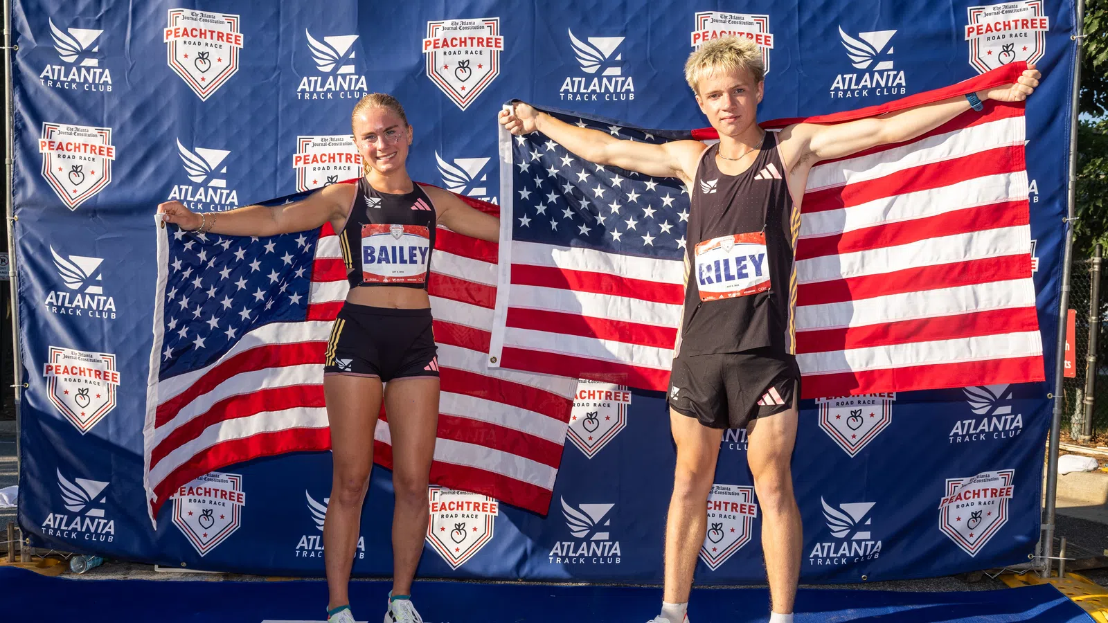 Two young athletes, Bailey and Riley, pose holding American flags at the Peachtree Road Race backdrop in Atlanta.