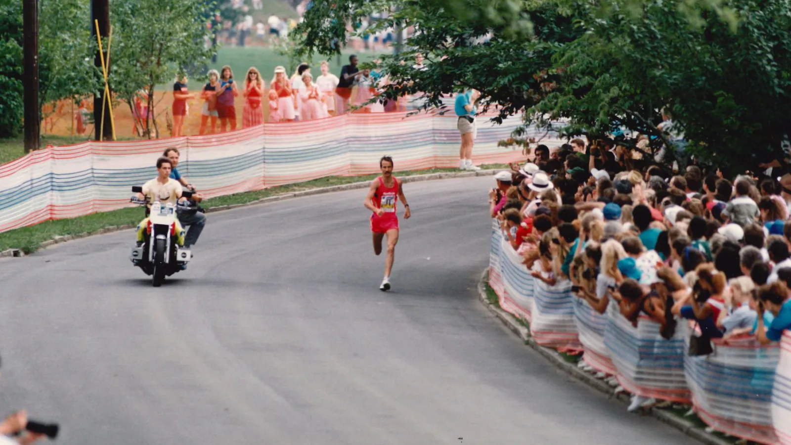 Runner in red competing on a curved road with crowd cheering behind safety barriers and a motorcycle escort.