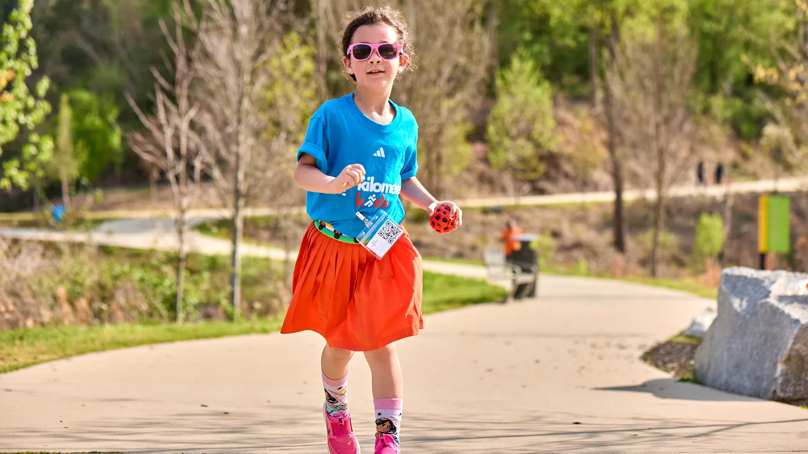 Young girl in blue shirt and orange skirt running outdoors on a sunny day with sunglasses and pink shoes.
