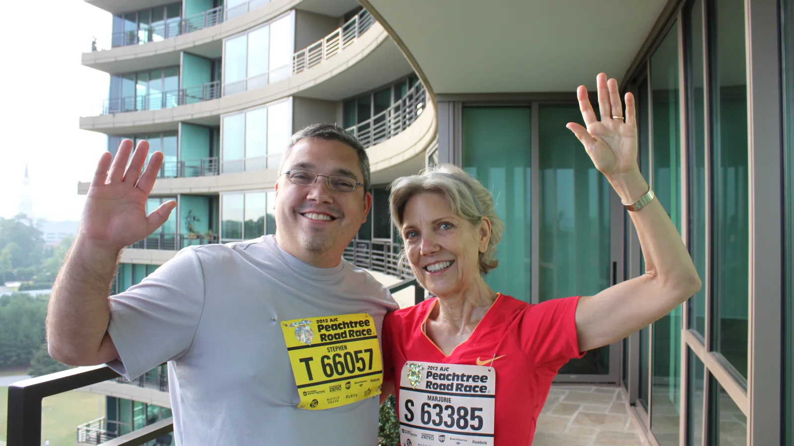 Man and woman with race bibs smiling and waving on a balcony before the Peachtree Road Race.