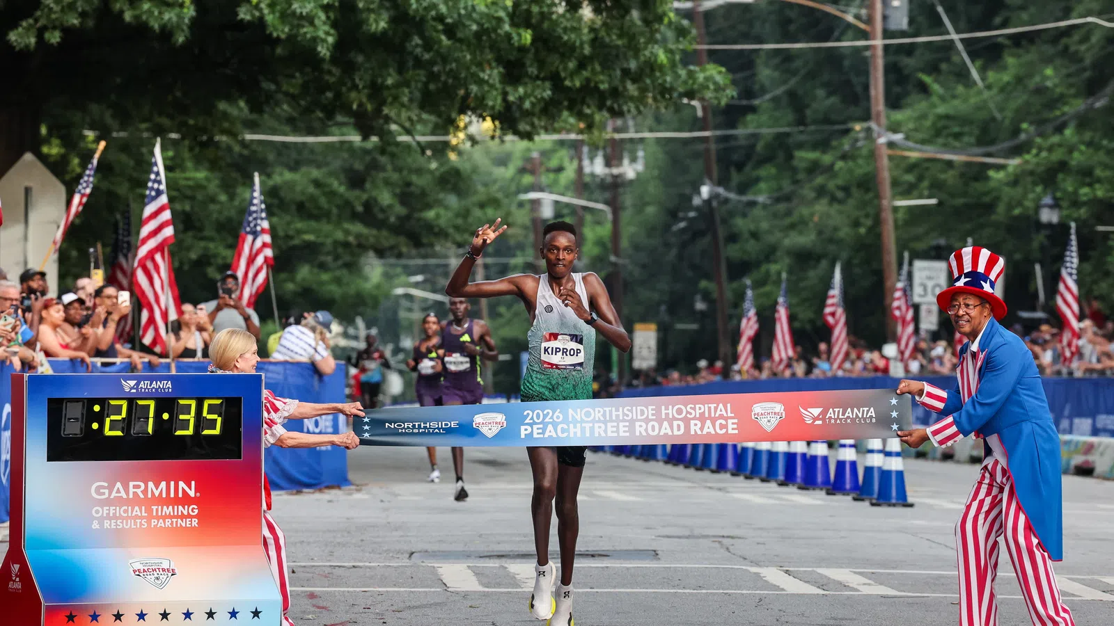 Runner Kiprop crossing the finish line of the 2026 Peachtree Road Race with a time of 27:35 in festive setting.