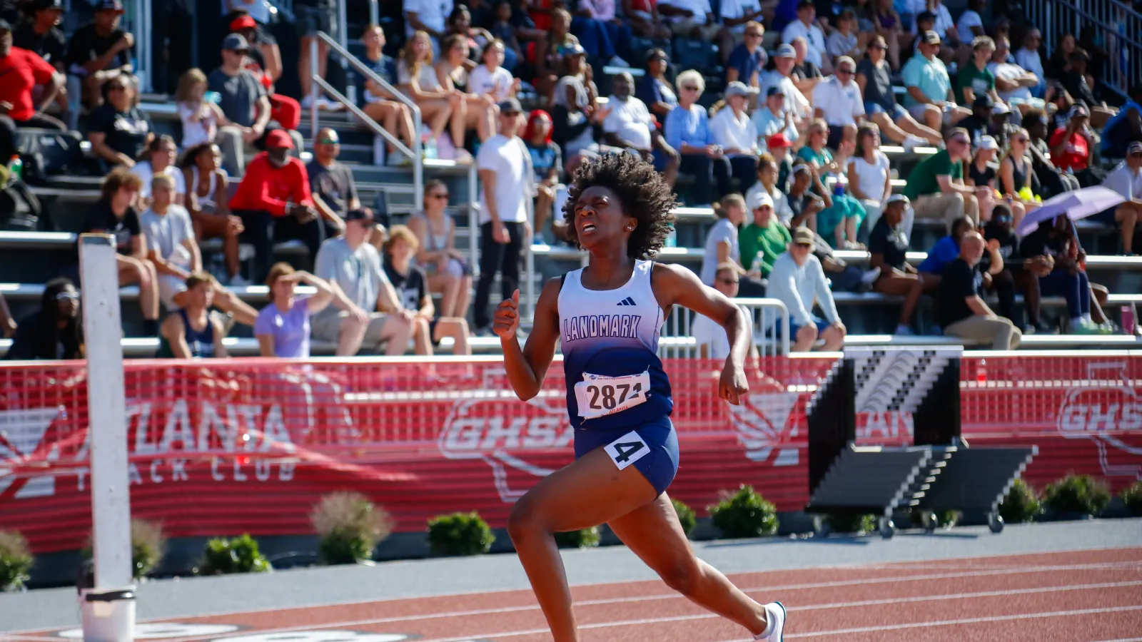 Female athlete sprinting on track during a crowded outdoor track and field event with spectators cheering.