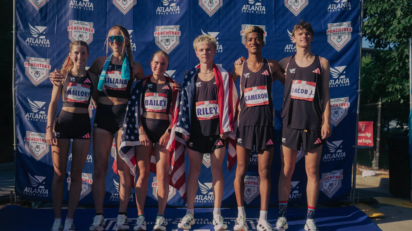 Six young runners posing with American flags at the Peachtree Road Race finish line backdrop in Atlanta.