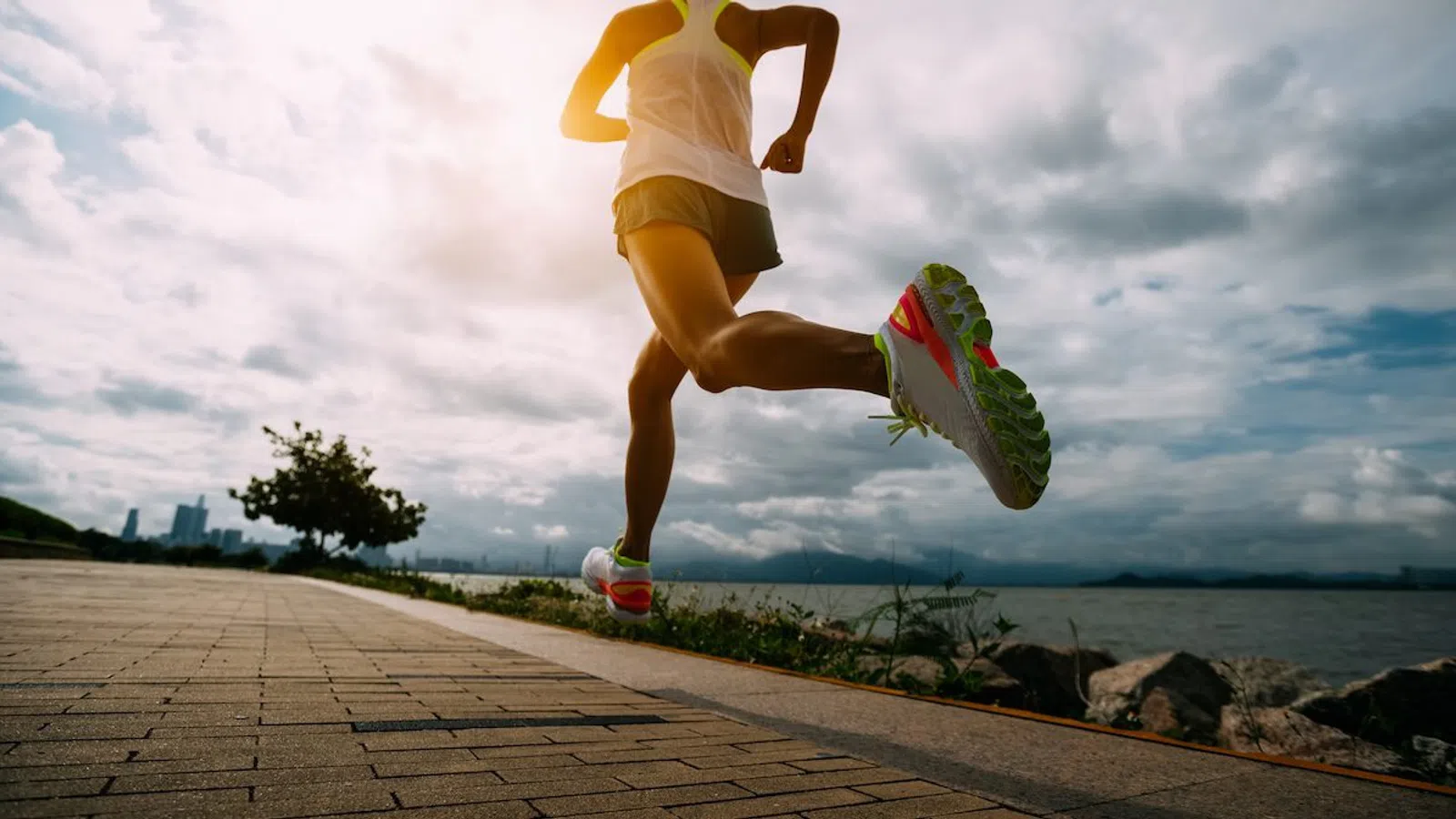 Runner jogging along a waterfront path under a cloudy sky with city skyline in the background.
