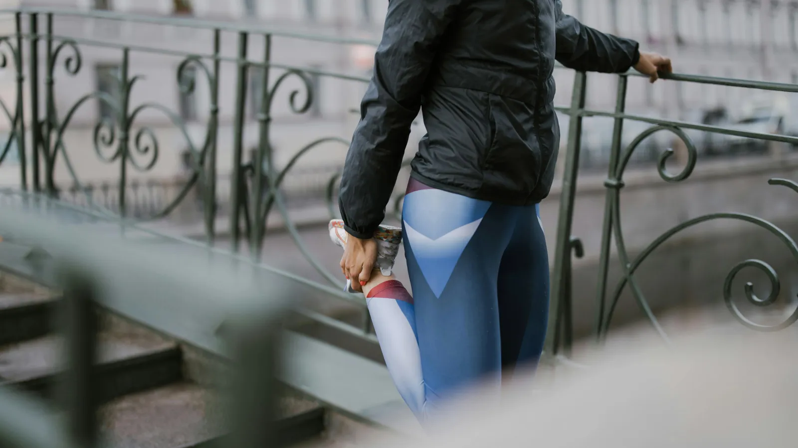 Person wearing a black jacket and colorful leggings stretches by holding one foot near a metal railing outdoors.