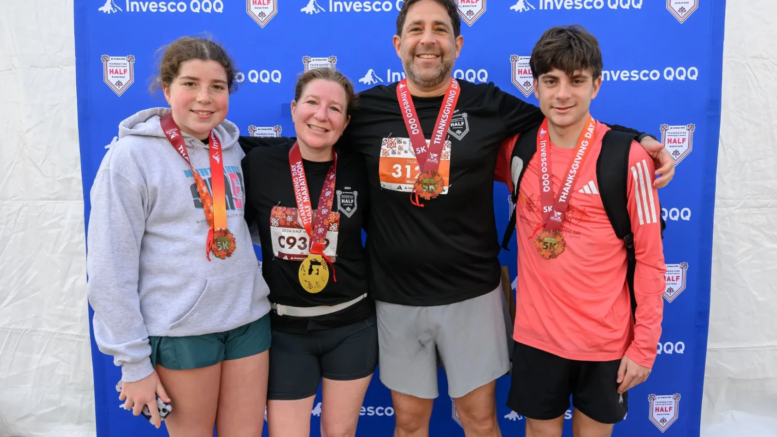 Four runners wearing medals stand smiling in front of an Invesco QQQ race backdrop after a 5K race.