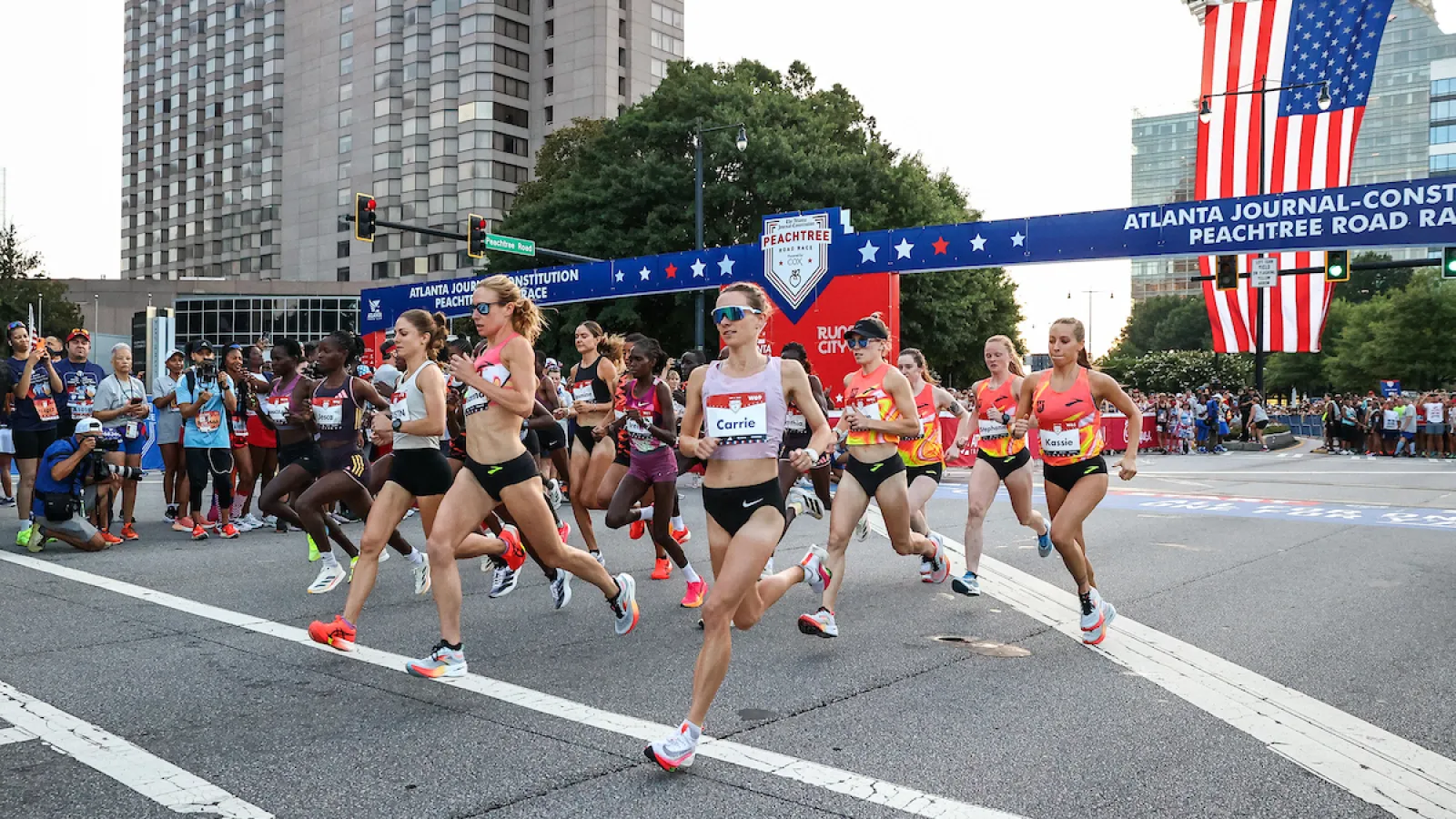 Female runners sprinting at the start of the Peachtree Road Race in Atlanta with American flag overhead