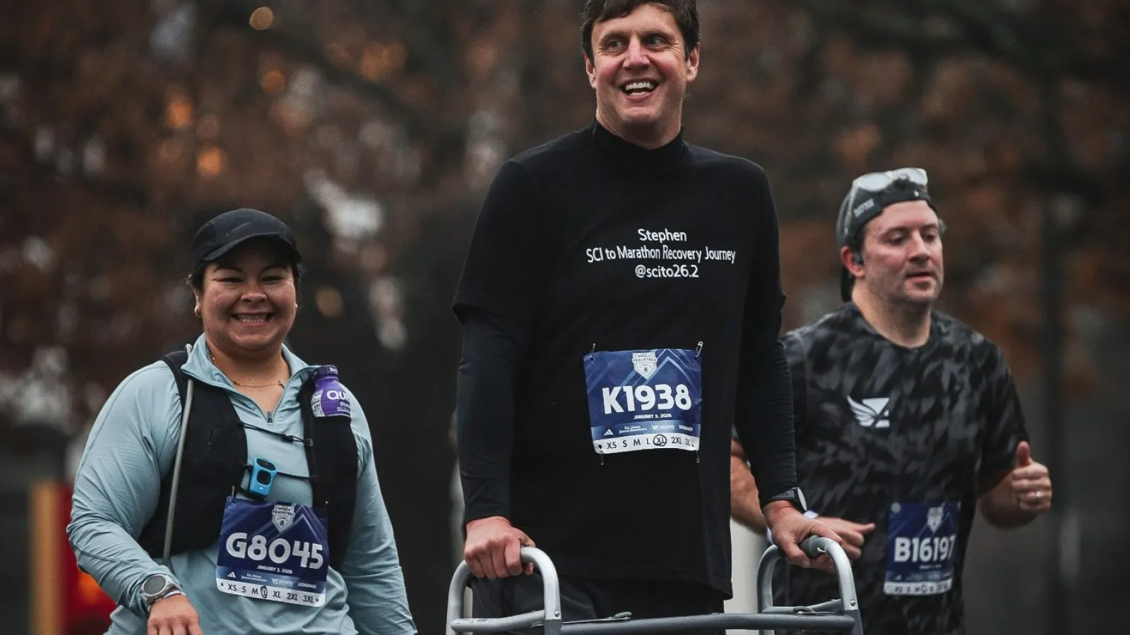 Three marathon runners, one using a walker, smiling and running together on a city street in autumn.