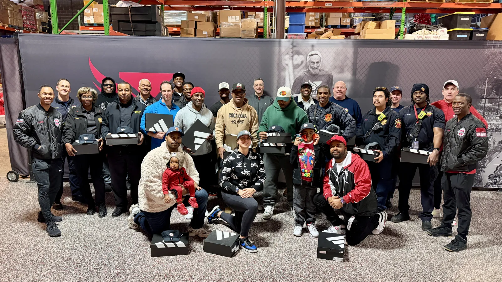 Group of diverse adults and children holding Adidas shoe boxes in a warehouse setting with shelves and boxes.