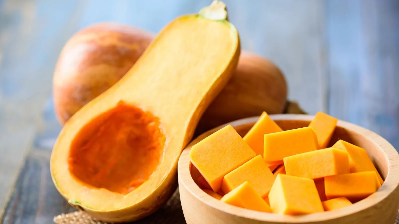 a bowl cut up butternut squash next to two halves of a whole squash, one with a hole carved out in the center