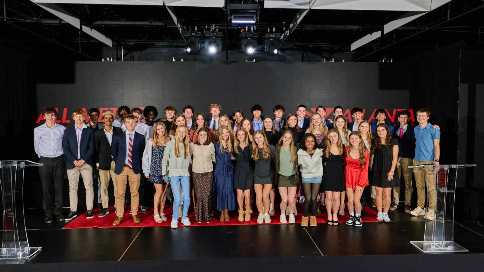 Large group of young adults posing on stage in smart casual attire under bright stage lights with a dark backdrop.