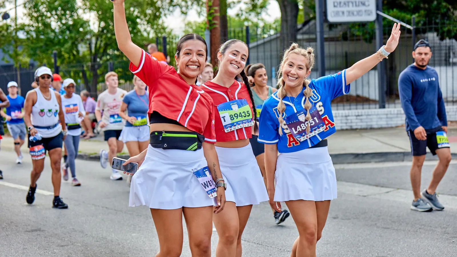 Three cheerful female runners in colorful outfits wave during a marathon event.
