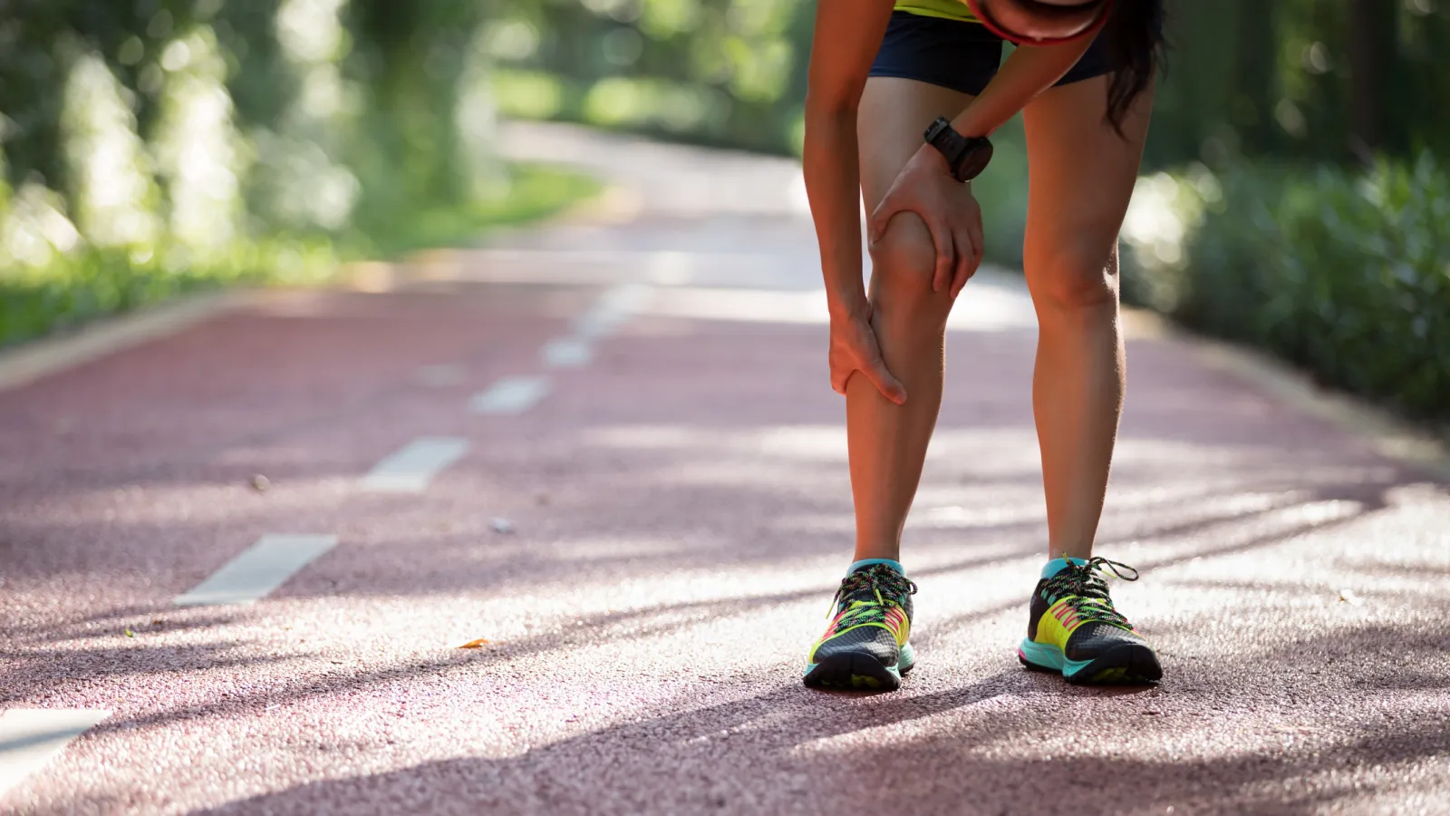 Runner holding knee in pain on a sunlit outdoor track surrounded by greenery during exercise.