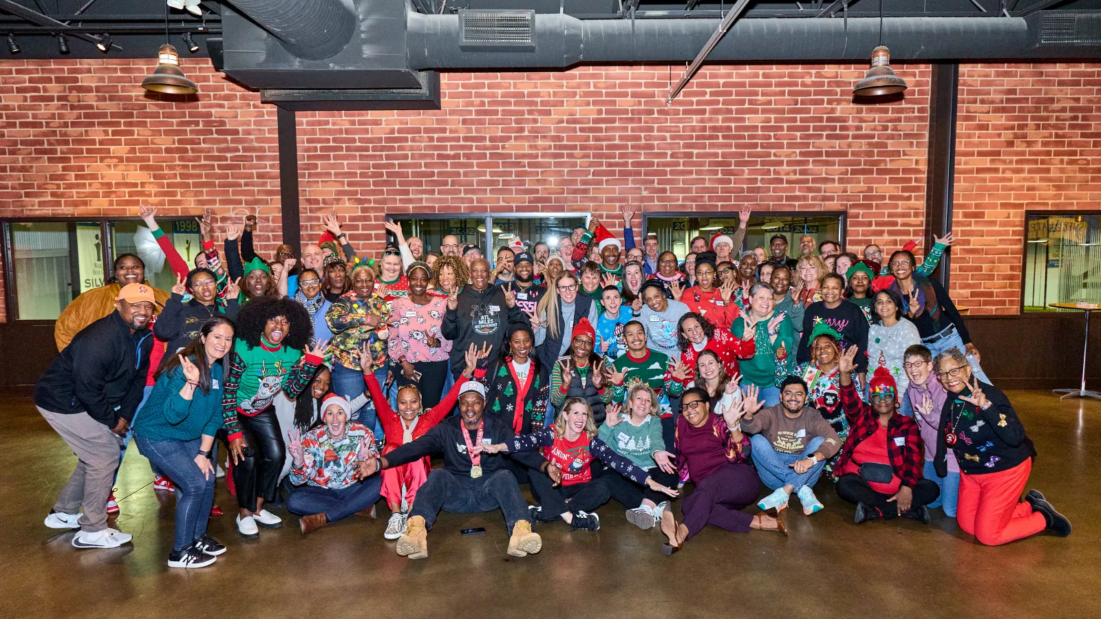 Large diverse group wearing festive holiday sweaters posing indoors against a brick wall and waving at the camera