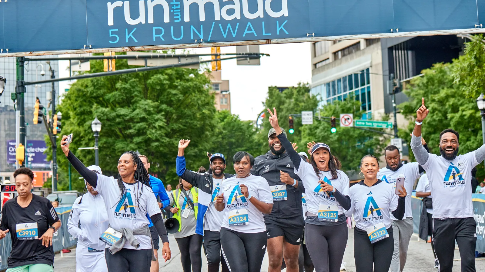 Group of diverse runners celebrating under Run with Maud 5K Run/Walk event banner in urban setting