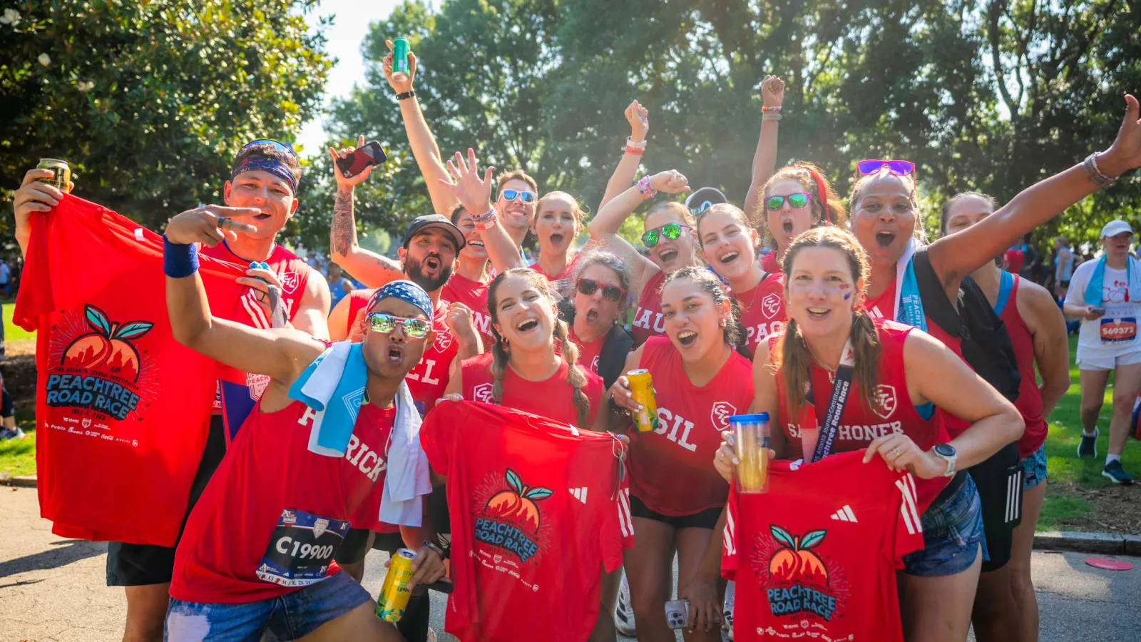 Group of joyful runners in red shirts holding Peachtree Road Race shirts celebrating outdoors in sunny park