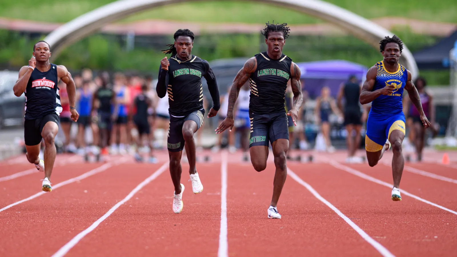 Four male sprinters competing on a red track during a daytime outdoor race with spectators blurred in the background