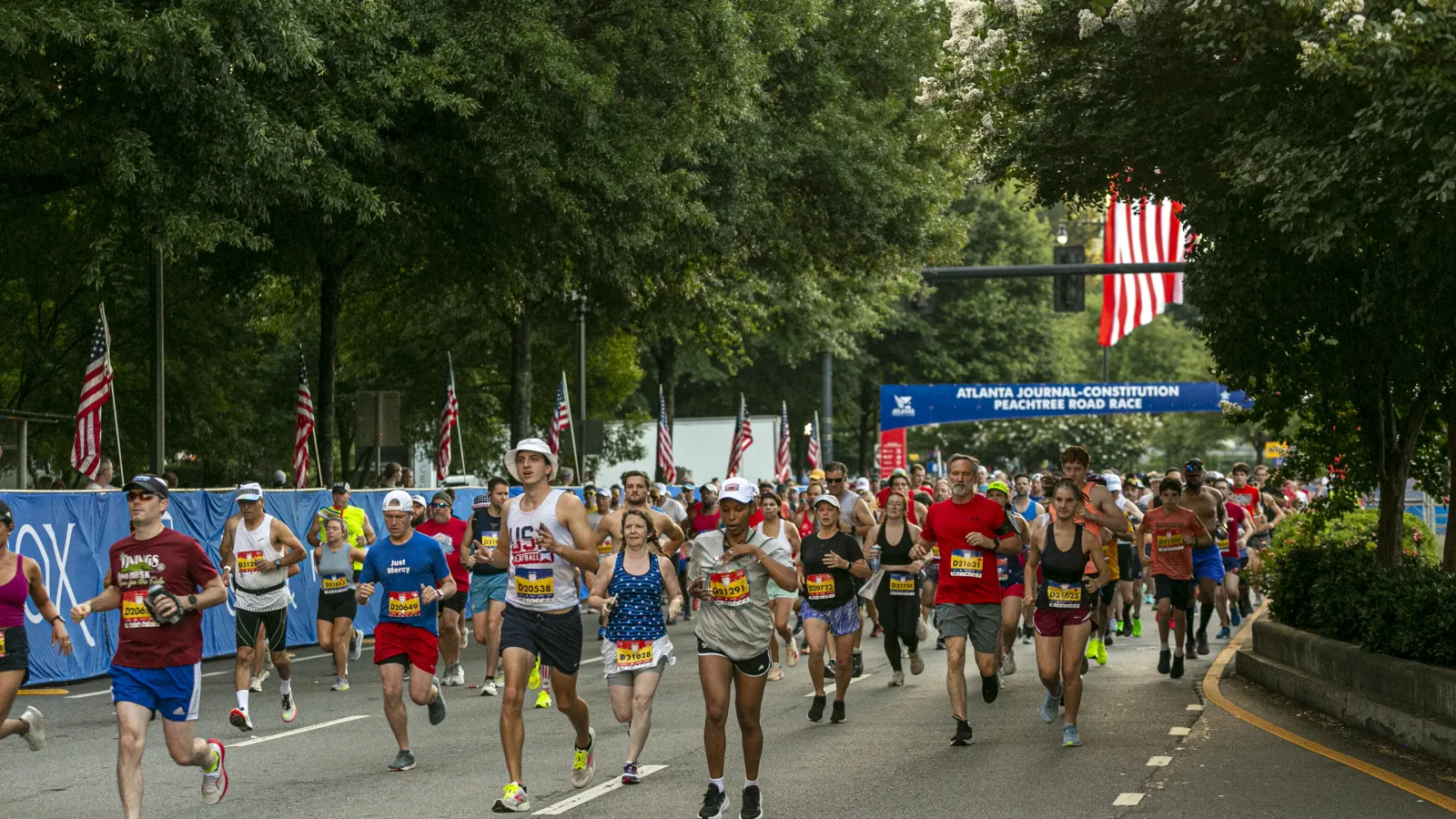 Runners participating in the Atlanta Journal-Constitution Peachtree Road Race on a tree-lined street with American flags.