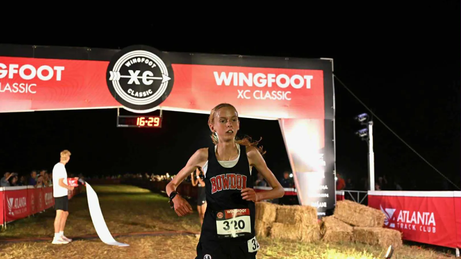 Young female runner in Bowdon uniform crosses finish line at night in Wingfoot XC Classic race event.