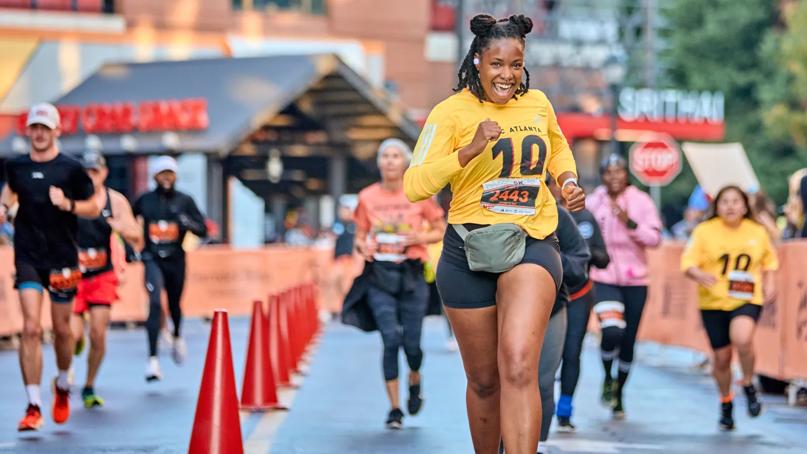Smiling woman in yellow shirt running in a city marathon with other participants behind her on a blocked street.