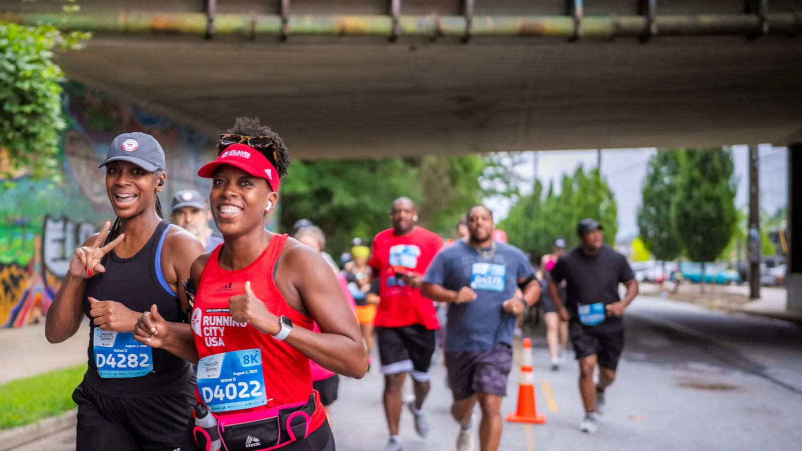 Runners smiling and jogging under Atlanta Beltline Lawton Street bridge during a city race event.