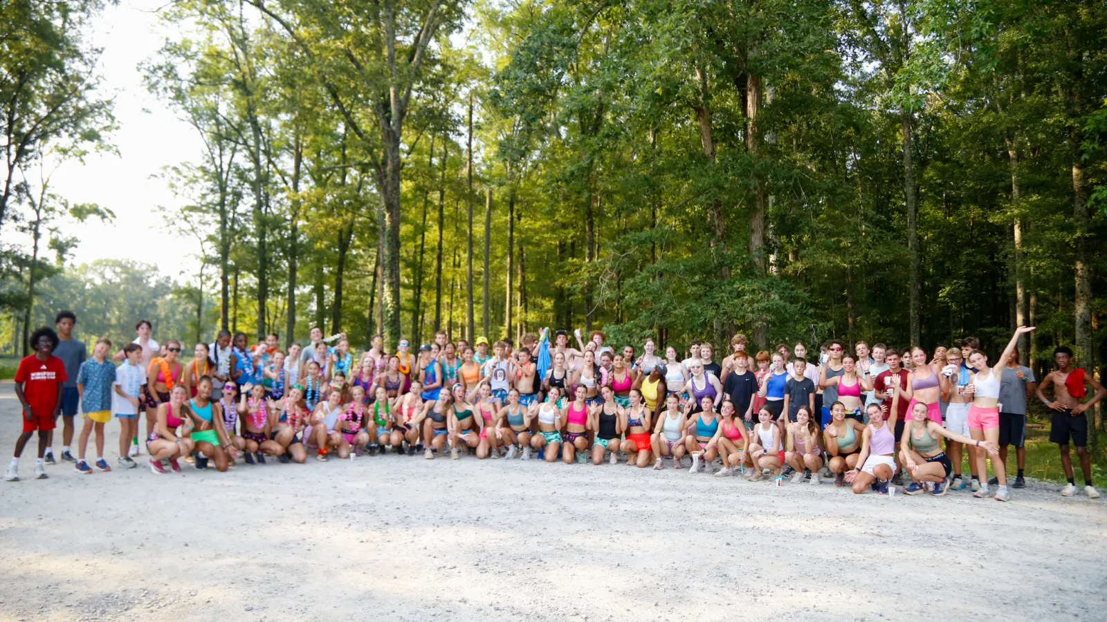 Large diverse group of people in athletic wear posing outdoors on a gravel area surrounded by tall trees.