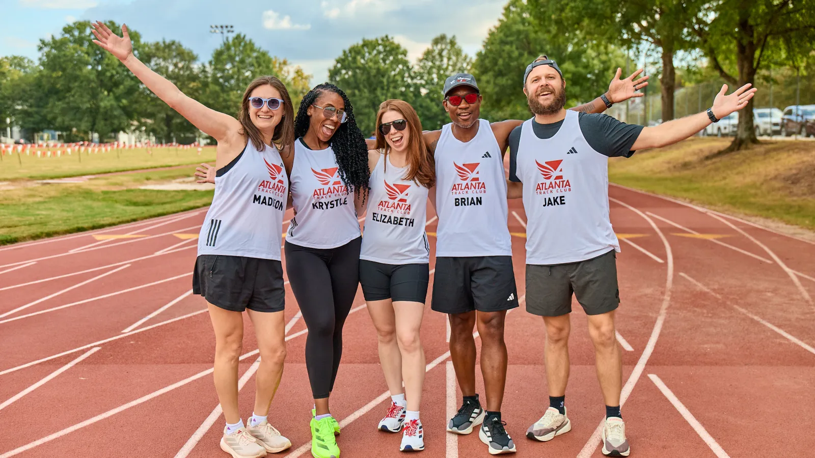 Five smiling runners wearing Atlanta Running Club shirts standing on a track with arms raised.