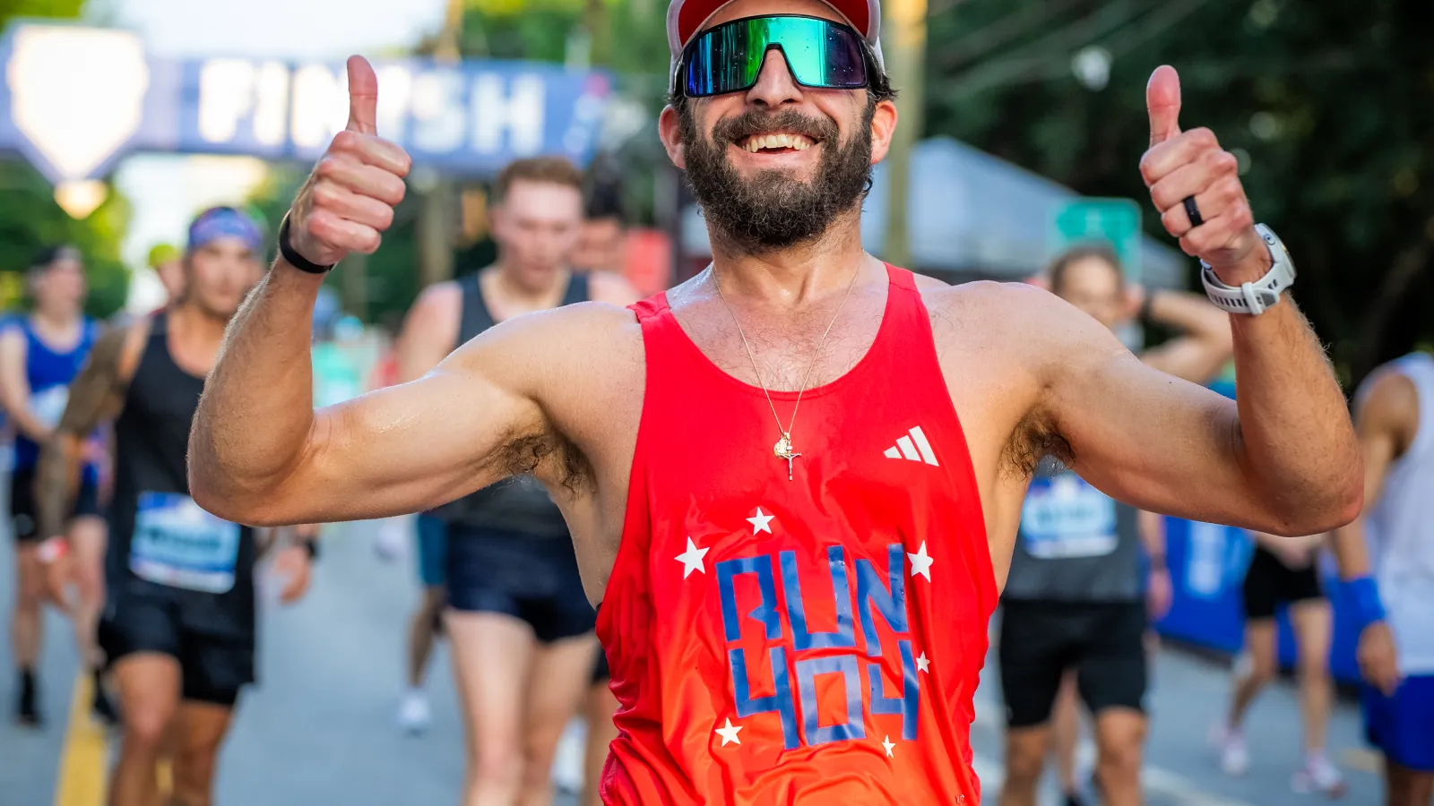 Smiling male runner in red tank top gives thumbs up near marathon finish line with runners behind him.