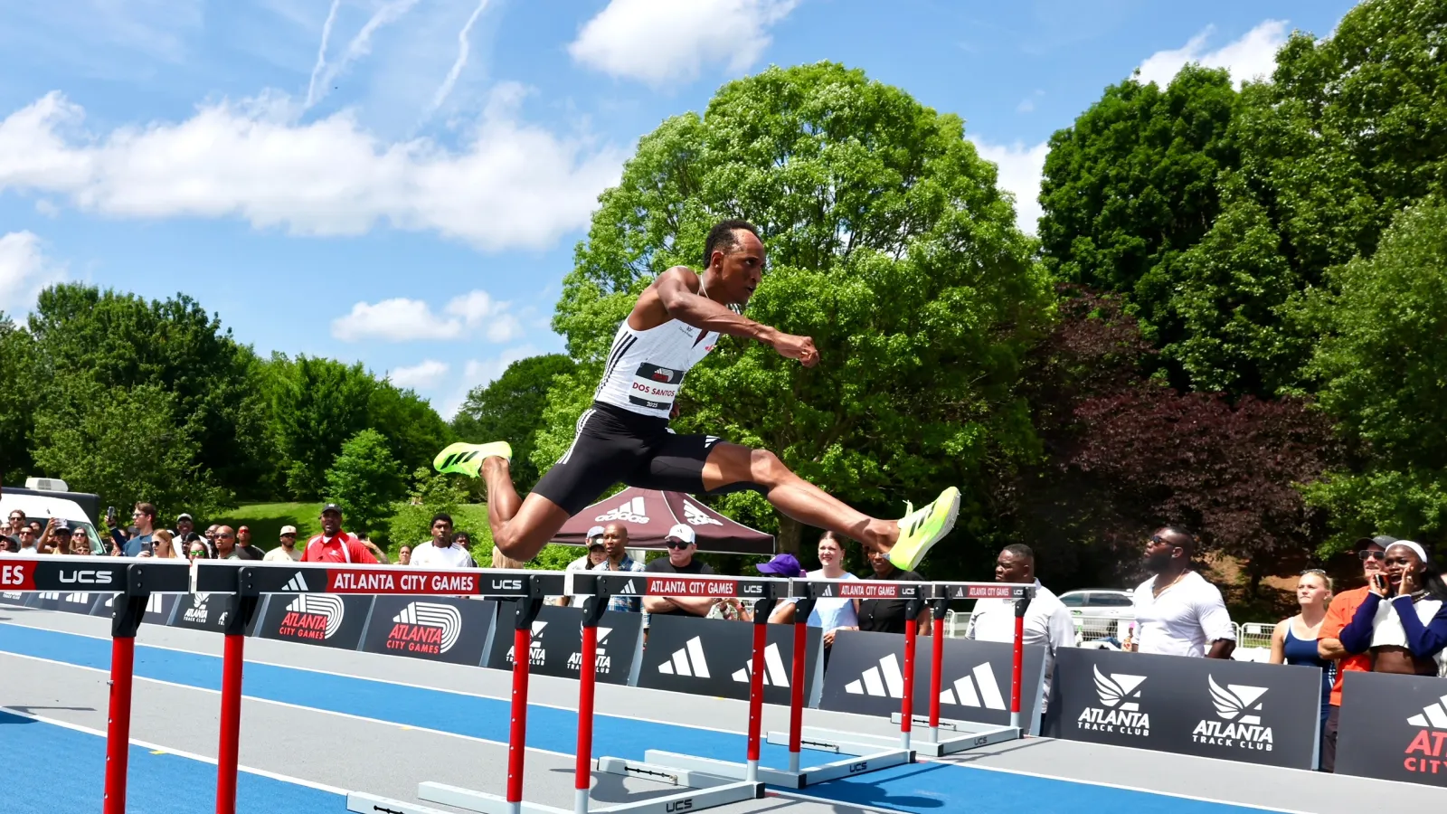 Athlete in mid-air clearing hurdle at Atlanta City Games with spectators and green trees in background.
