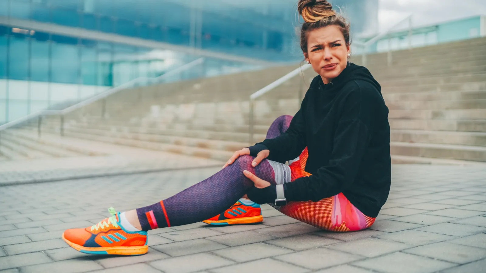 Young woman in colorful athletic wear sitting on urban stairs holding her knee with a pained expression