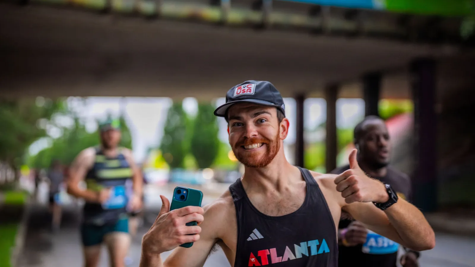 Smiling runner wearing an Atlanta tank top gives thumbs up during a race under a bridge with other runners in the background