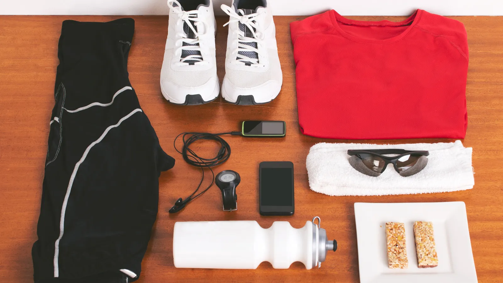 Flat lay of workout gear including shoes, black pants, red shirt, water bottle, snacks, sunglasses, towel, and fitness gadgets.