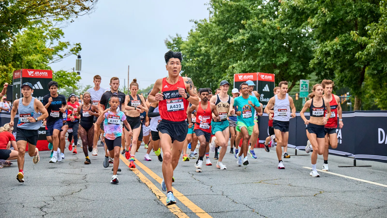 Runners participating in a road race event wearing bibs, running on a street lined with trees and banners.