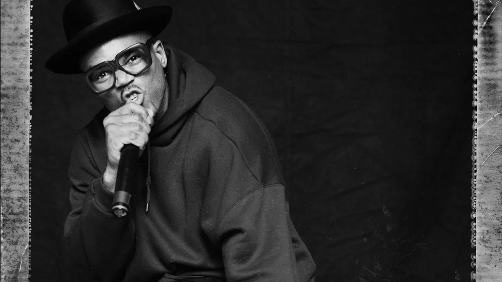 Black and white photo of a rapper in glasses and hat holding a microphone with DMC logo in corner.