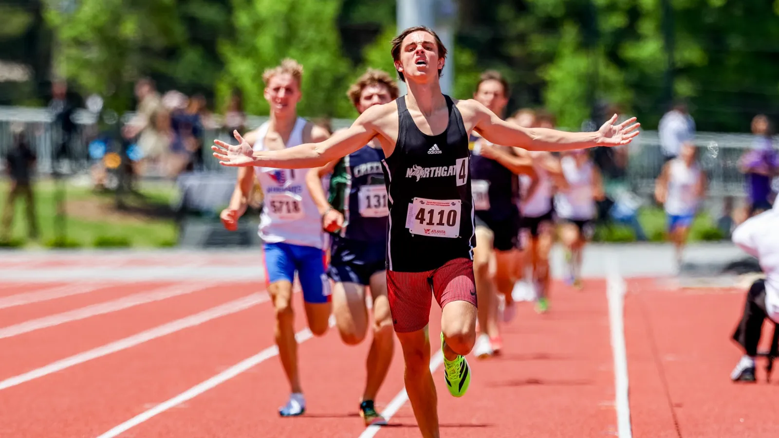 Young male athlete crosses the finish line with arms spread wide leading a track race on a red running track.