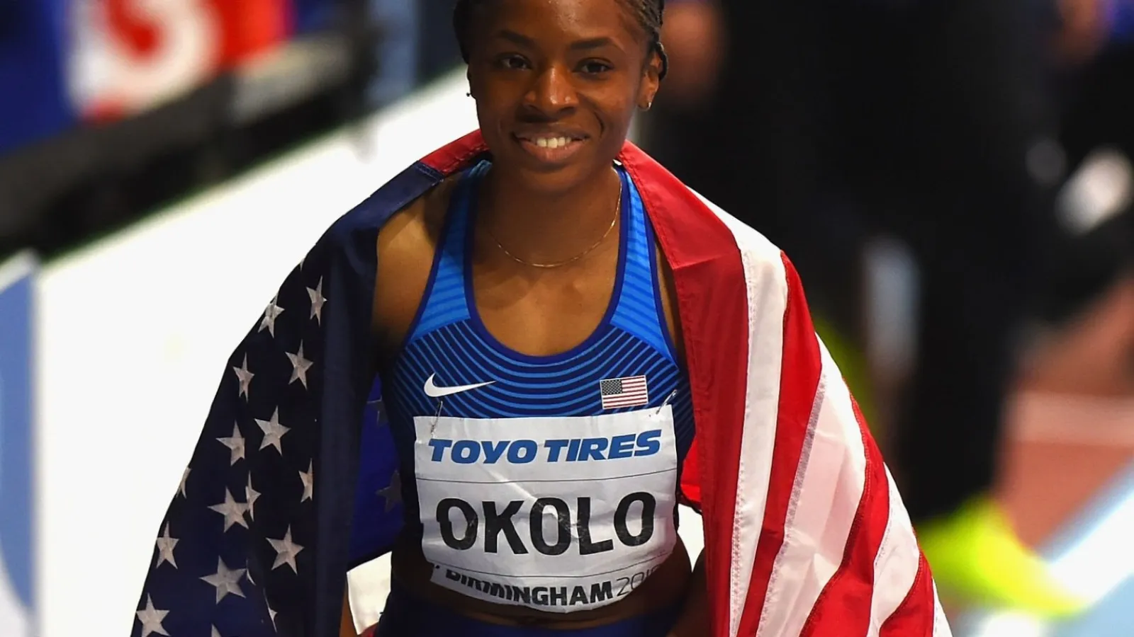 Courtney Okolo in a blue and white track suit with a flag behind on her shoulders