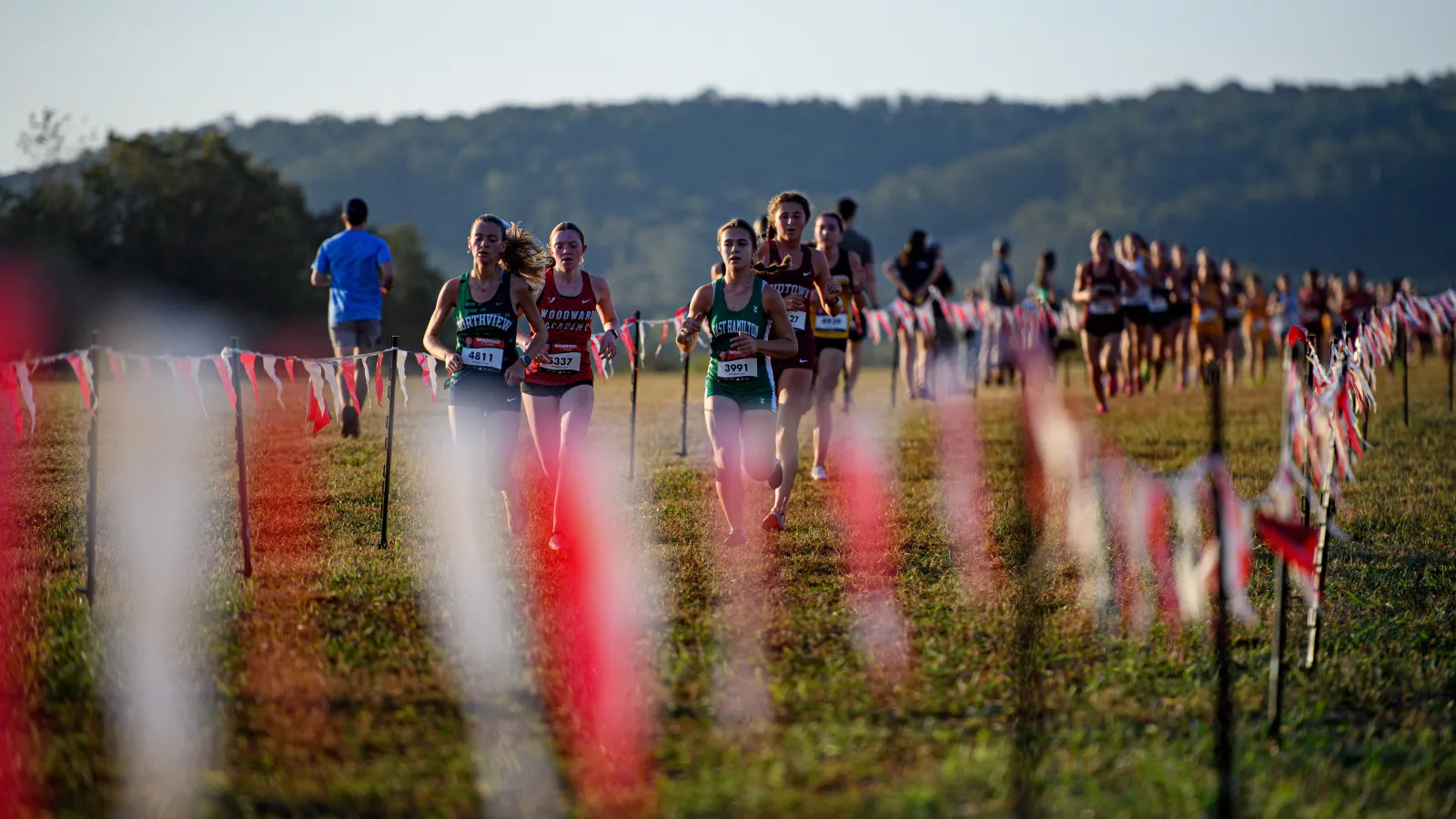 Female cross-country runners competing on a grassy course marked with red and white flags under a clear sky.
